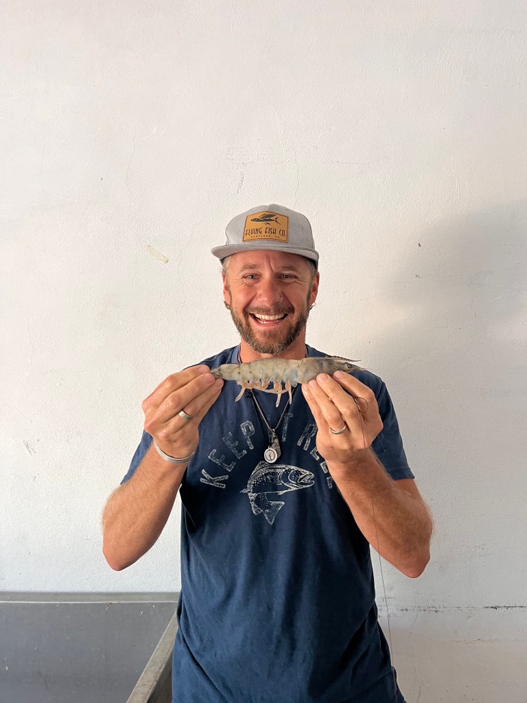 A smiling man holds a large shrimp in front of a plain white background, wearing a gray cap and a blue t-shirt.