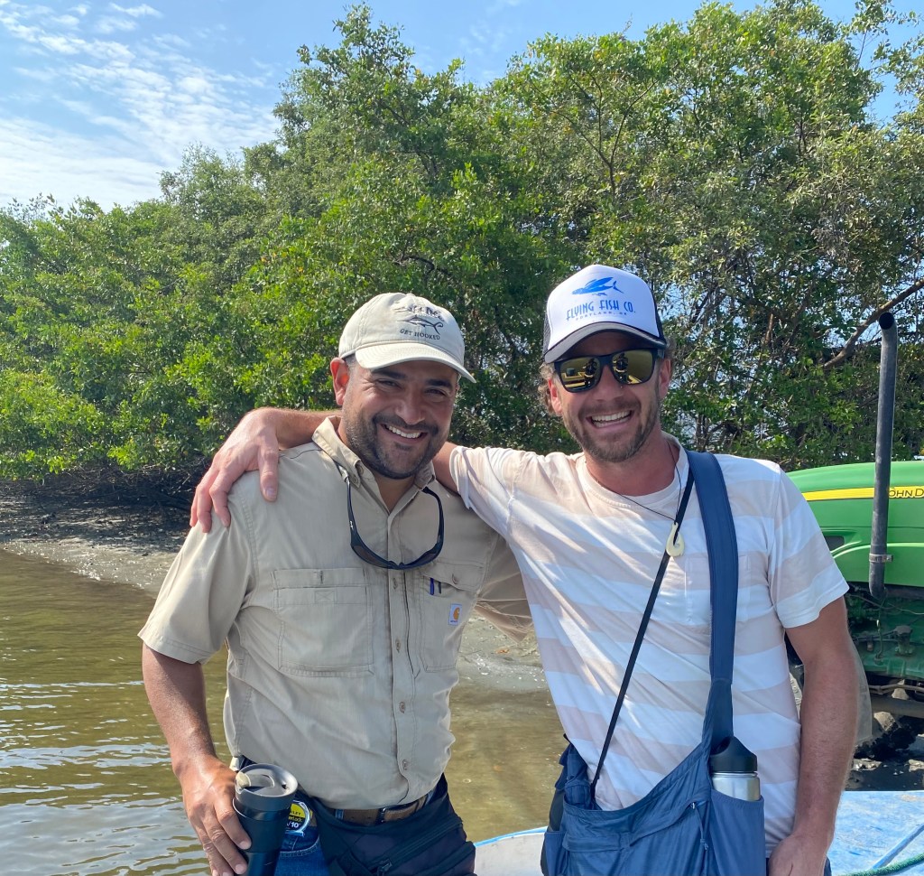 Two men posing together on a boat near mangroves, one wearing a light-colored shirt and a cap, the other in a striped shirt and sunglasses, with a green tractor in the background.