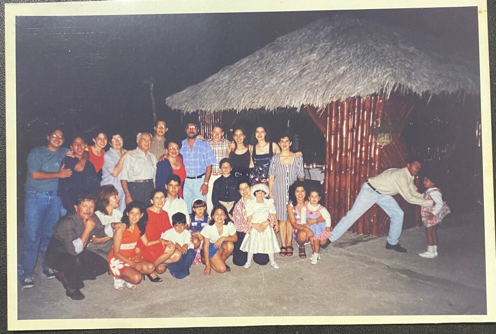 A group photo of a large family gathering outdoors, with a thatched-roof structure in the background. There are adults and children posing together, smiling and some playfully interacting with each other.